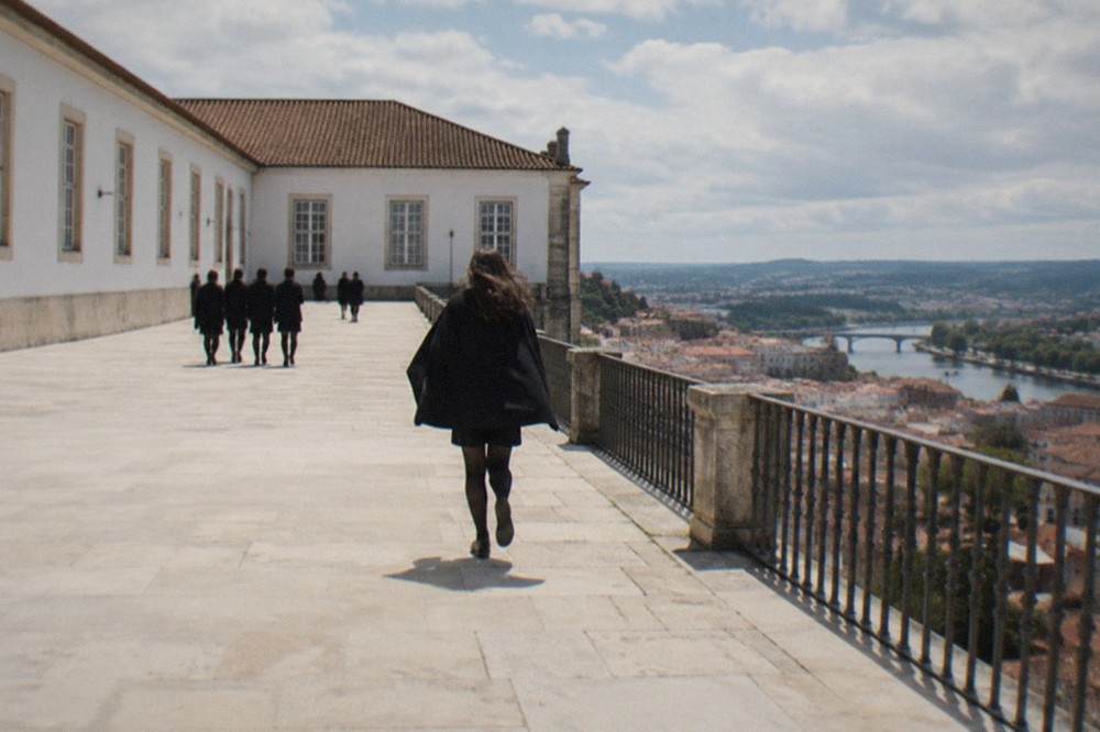 Students in dark capes crossing a cobbled square below the University of Coimbra
