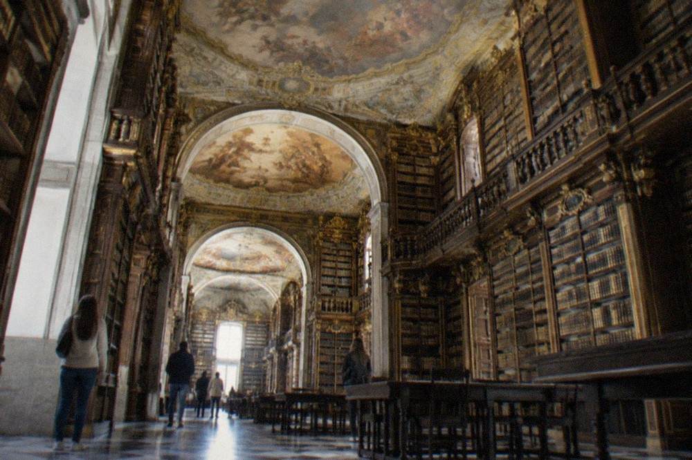 Visitors viewing gilded bookshelves in a baroque library room with painted ceiling