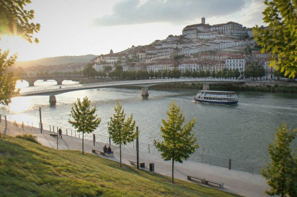 A small cruise boat moving along the Mondego with the arched bridge and old town buildings behind it