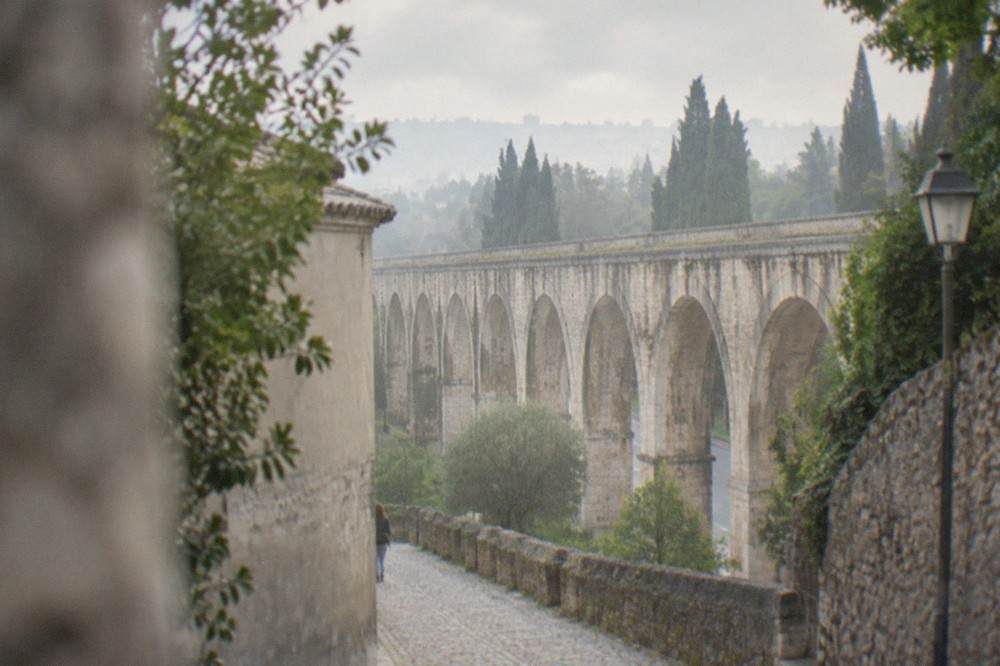 A cobbled lane in Coimbra climbing past stone walls with a line of aqueduct arches visible through trees