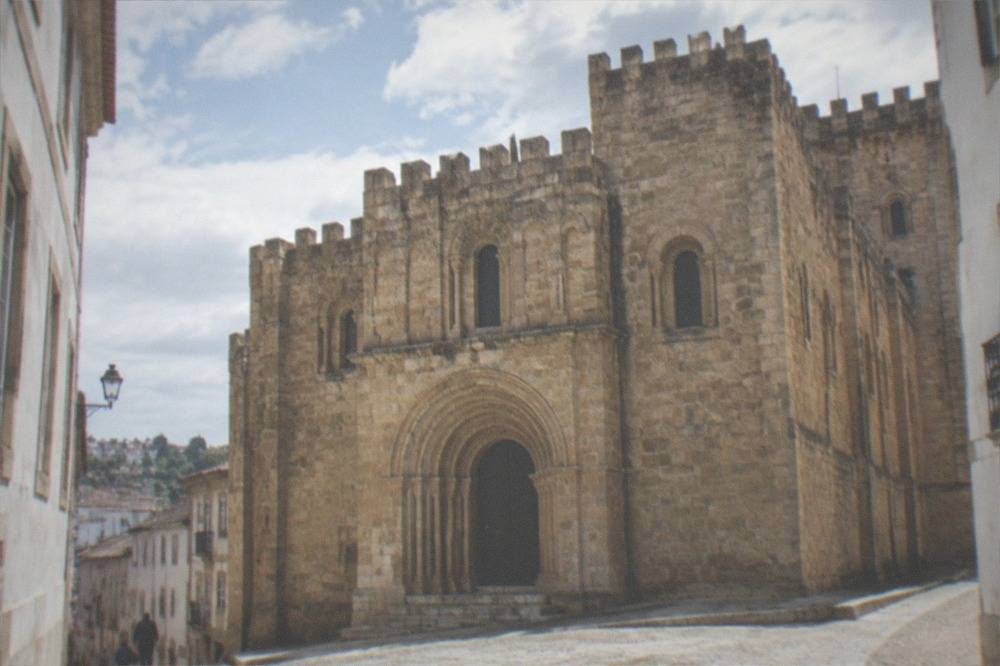 A Romanesque cathedral with crenellated roofline seen from a narrow stone street in Coimbra