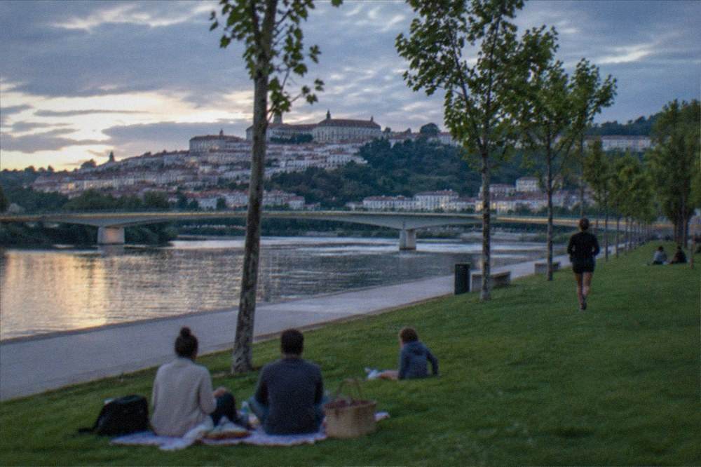 Family picnicking on riverside grass with the Mondego and a bridge in the background