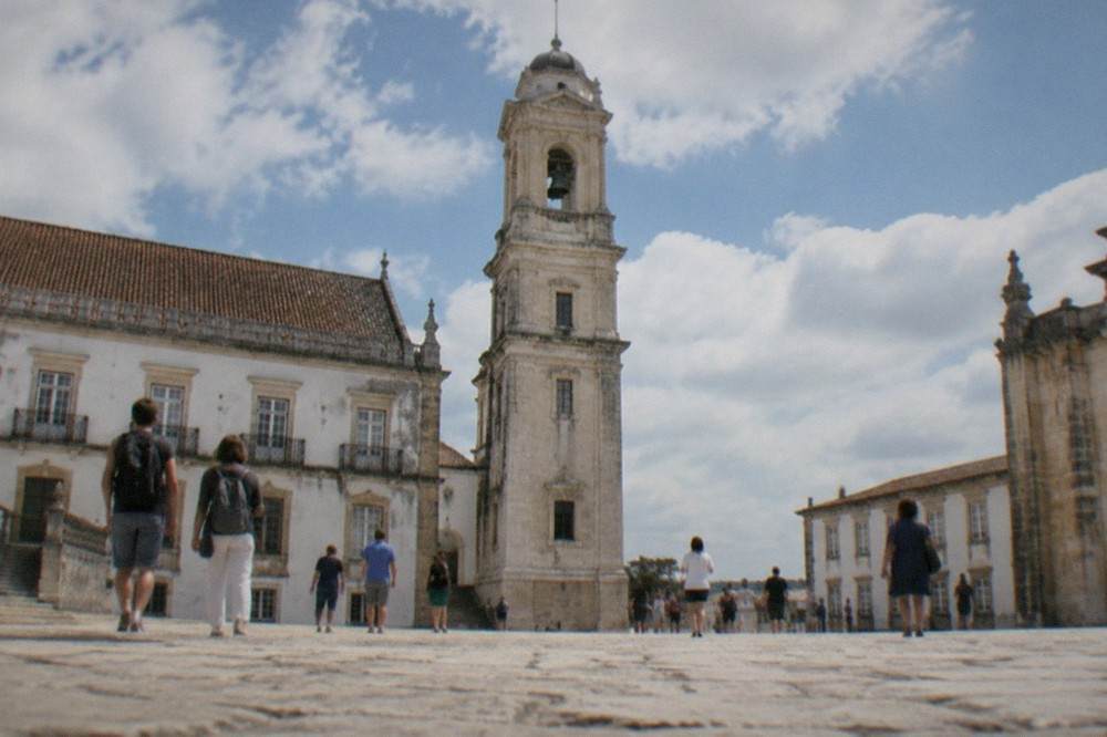 Tourists walking through the stone courtyard of the Paço das Escolas with the clock tower above