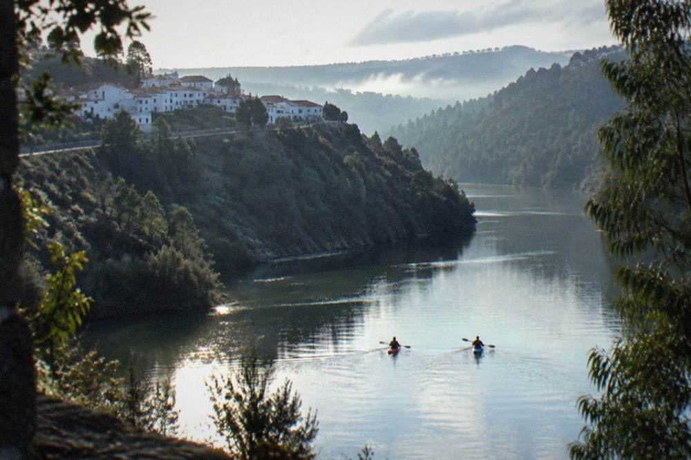 Two kayakers paddling on wide flat water in early morning light, mist over far hills