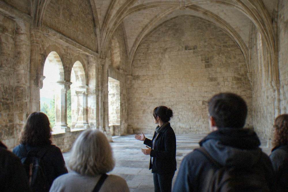 A guide speaking to a small group of visitors in a large stone chapter house with a vaulted ceiling