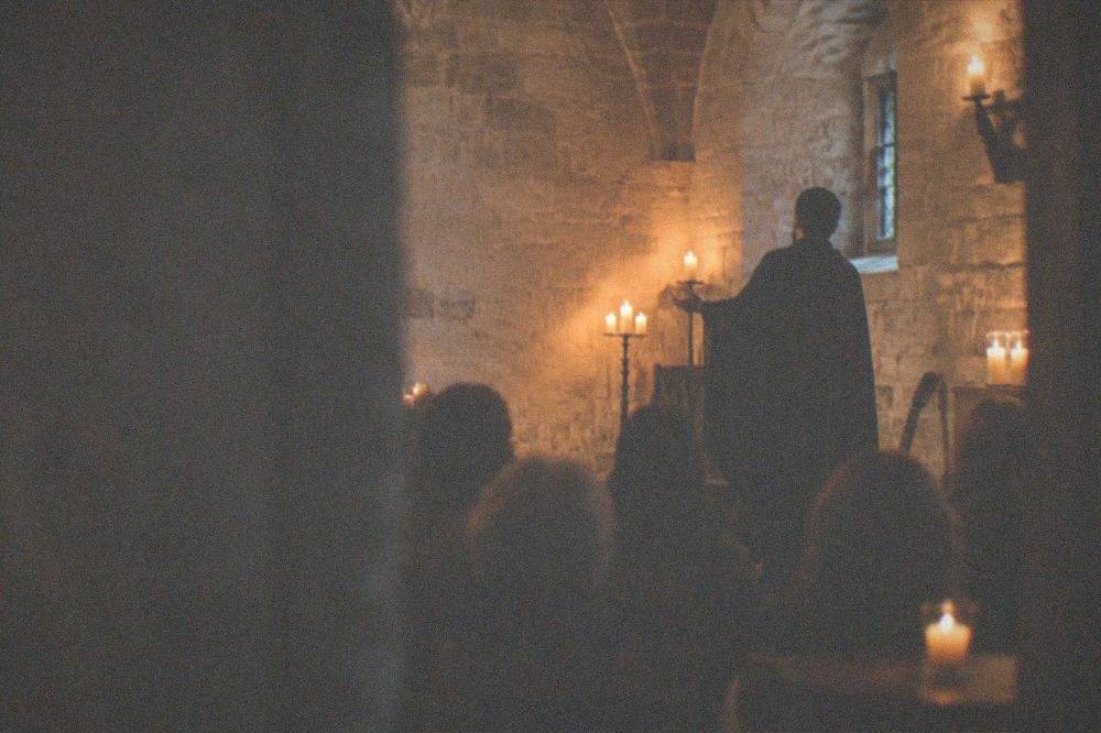 A singer in a dark cape performing to a small seated audience in a candlelit stone room