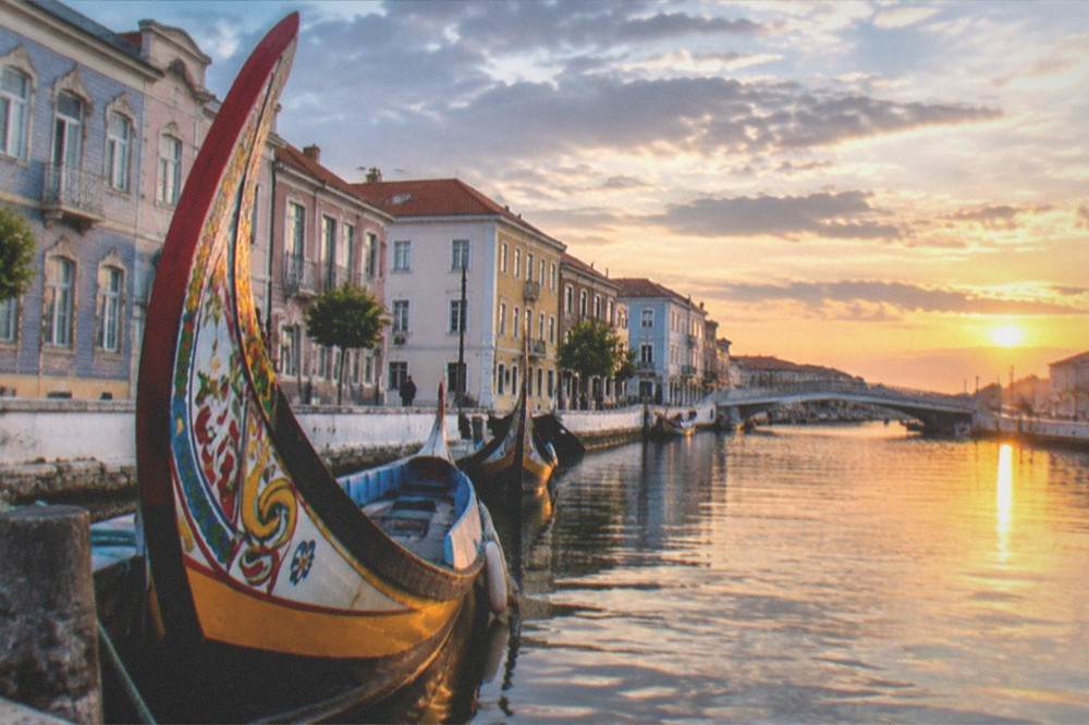 A canal in a Portuguese lagoon city at sunset with small boats and pastel coloured buildings along the water