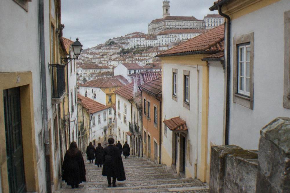 Students in black capes walking through a narrow Coimbra street lined with old buildings