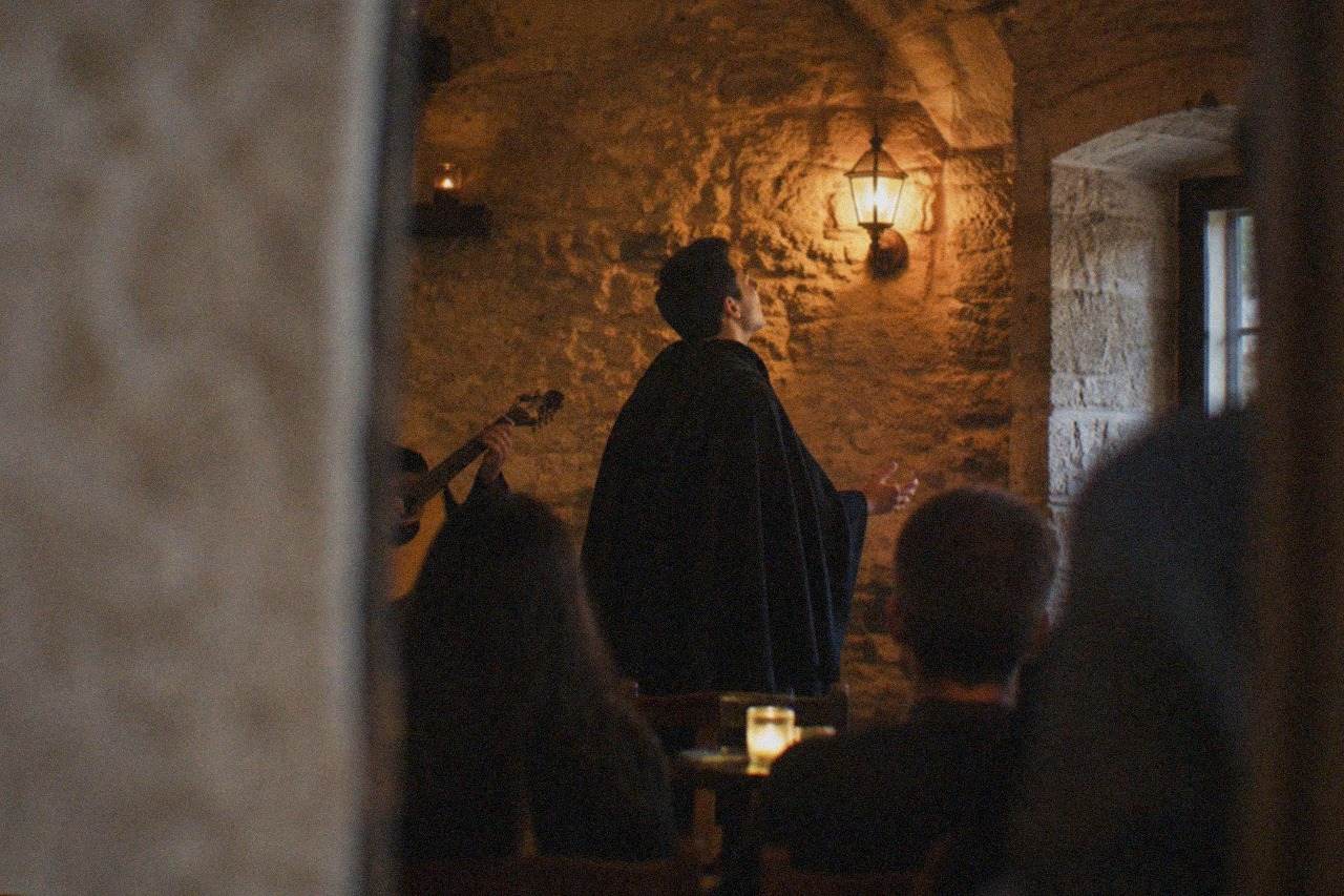 A singer in a dark academic cape performing under warm lamplight in a small stone-walled room in Coimbra