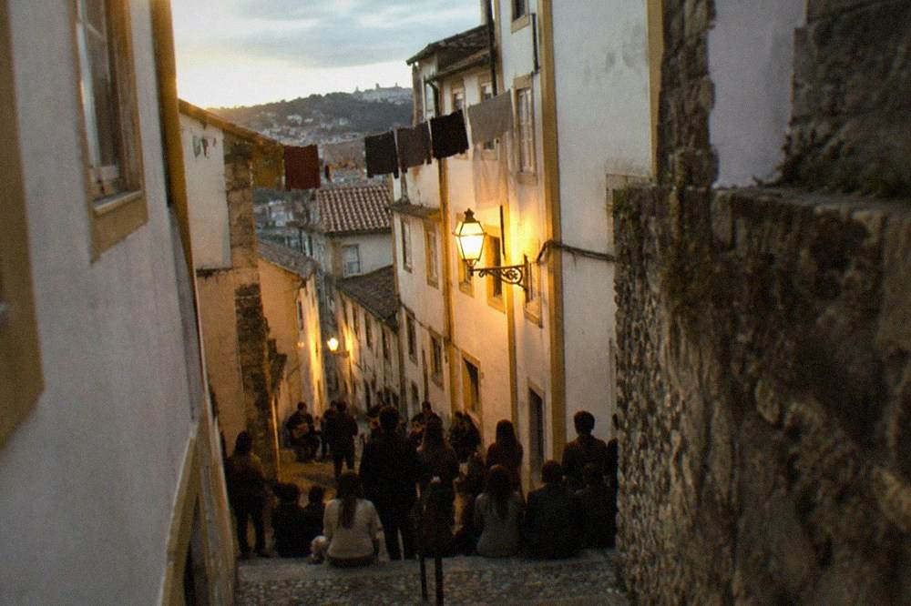 A narrow lamplit street in Coimbra's old town at night with listeners gathered around a small group of singers