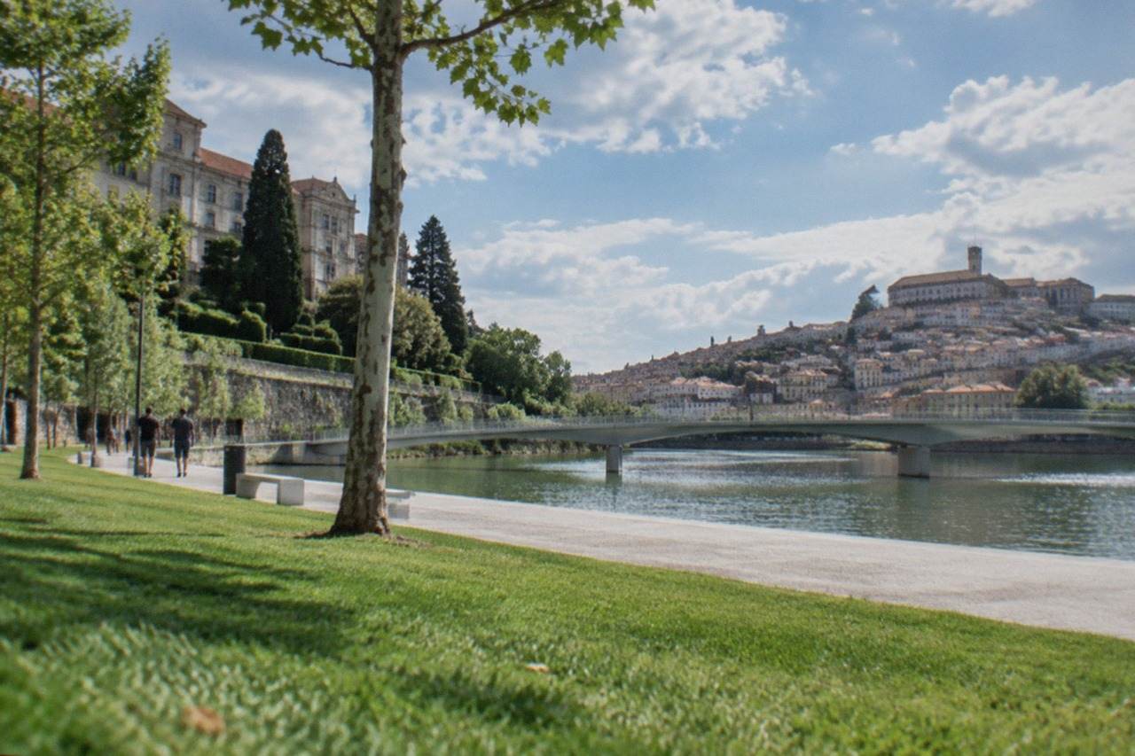A historic stone building with manicured gardens and tall trees beside the River Mondego in Coimbra