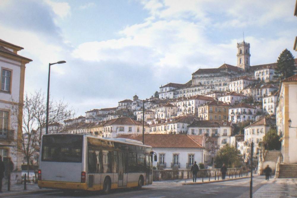 A city bus stopped on a street in central Coimbra with the old town rising behind it.