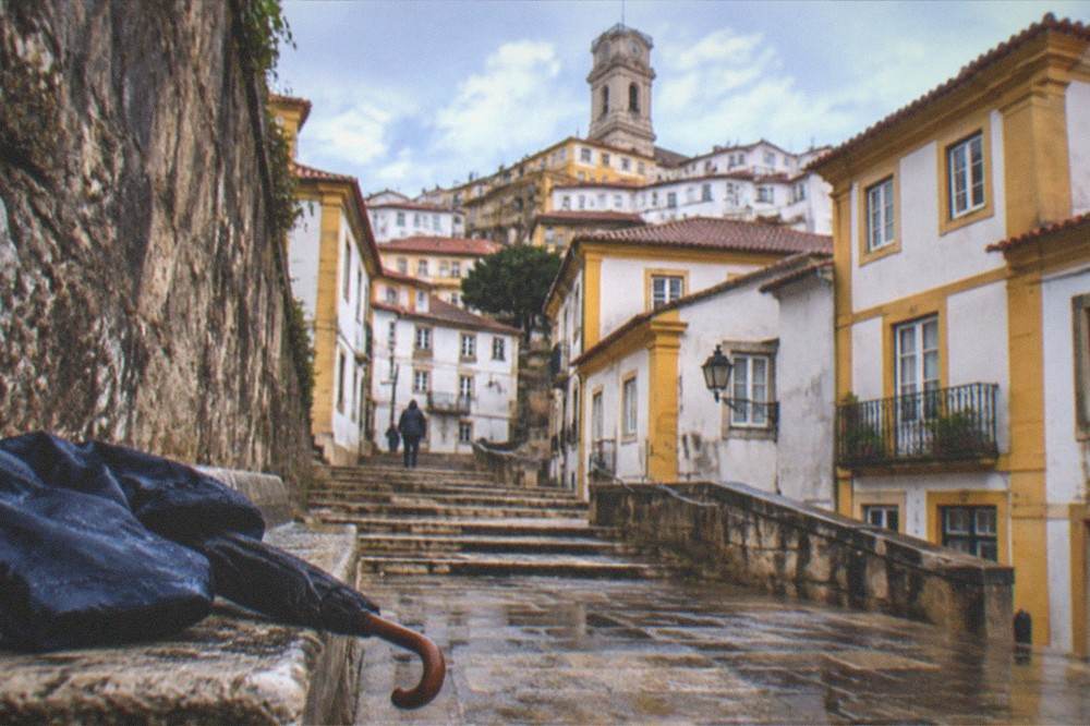 Umbrella and wet rain jacket resting on a stone bench beside a rain-flecked medieval wall in Coimbra