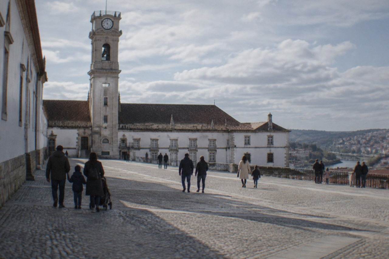 Families walking on a cobbled square below the tower of the University of Coimbra on a bright day