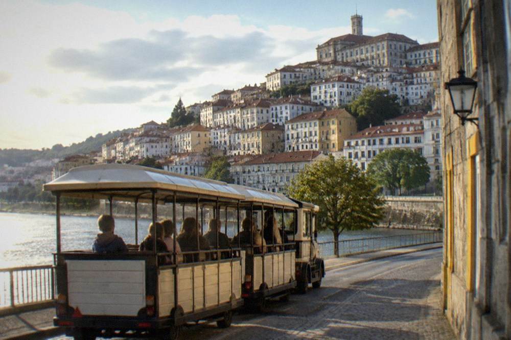 Children riding a small open-sided tourist train along a riverside road with the city hill behind